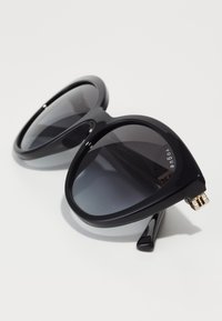 Ochelari de soare supradimensionați, negri, cu o ramă din plastic neted și lentile întunecate cu gradient. Prezintă un accent auriu pe balama templului. Formă unghiulară, tip cat-eye.