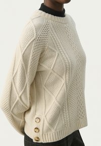 Cremfarvet strikket sweater med et tekstureret design med diamantmønstre, ribbet halsudskæring og tre træknapper på siden.