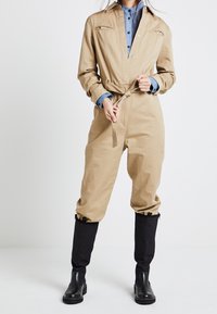 Khaki overall s úzkým pasem, dlouhými rukávy, náprsními kapsami a černými džínovými manžetami. Doplňuje se černými kotníkovými botami.