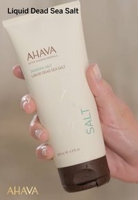 AHAVA Sale di Terra Morta Liquida in un tubetto bianco e marrone, etichettato in turchese, tenuto in una mano con finitura lucida e gocce visibili.
