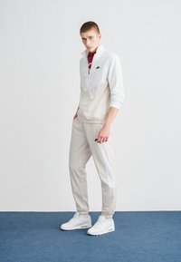 Chándal gris claro de Nike con chaqueta con cremallera que tiene la mitad superior blanca y la mitad inferior gris. Pantalones grises a juego y zapatillas blancas.