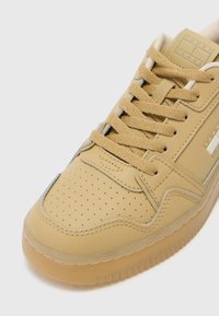 Tan leather sneaker with perforated toe, textured laces, and a rubber sole. Features a logo tag and subtle accent stitching.