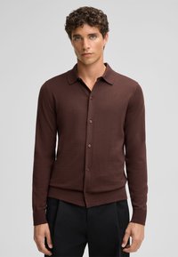 Camicia button-up a maniche lunghe di colore marrone scuro con colletto, tessuto texturizzato e bottoni marroni abbinati. Indossata con pantaloni neri.