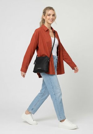 Frau trägt rostfarbenes Hemd, weißes Oberteil, hellblaue Jeans, weiße Turnschuhe und schwarze Umhängetasche, geht lächelnd vor einfachem Hintergrund.