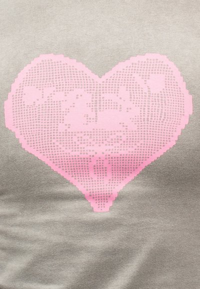 Ροζ pixel καρδιά με το ακρώνυμο "ZAZA" και μικρότερο, ακατανόητο κείμενο στο εσωτερικό, πάνω σε γκρι υφασμάτινο φόντο.