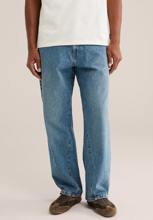 Persona con vaqueros azules holgados, zapatillas marrones y una camisa blanca, de pie sobre un fondo claro y liso.