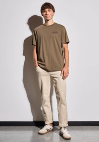 Olivově zelené bavlněné tričko s krátkým rukávem a potiskem na hrudi, zkombinované s béžovými bavlněnými kalhotami a bílými teniskami s hnědými akcenty.