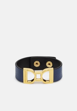 Marineblåt læderarmbånd med sløjfeformet lås i guldfarvet metal, graveret med "COCCINELLE" og to trykknapper.