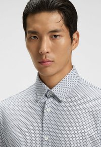 White button-up shirt featuring a navy geometric pattern, with a standard collar and smooth texture. Buttons are round and light-colored.