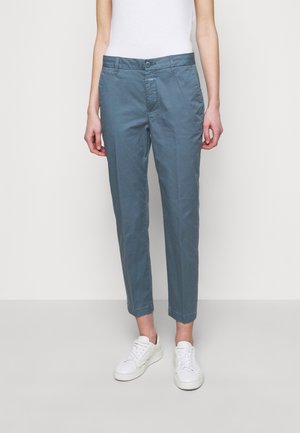 Femme portant un pantalon droit bleu raccourci et des baskets blanches, debout devant un fond clair uni.