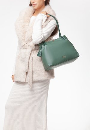 Donna con abito lavorato a maglia color crema e gilet di pelliccia che tiene una grande borsa in pelle verde sulla spalla.
