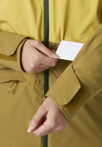 Olivgrüne und gelbe leichte Jacke mit Reißverschluss vorne, Velcro-Manschette und einer weißen Karte, die in eine Einschubtasche gesteckt wird.