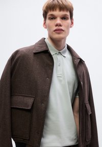 Καφέ σακάκι με ψαροκόκαλο μοτίβο, γιακά, κουμπιά μπροστά και δύο τσέπες, φορεμένο πάνω από ένα ανοιχτό πράσινο πόλο με μικρό λογότυπο.