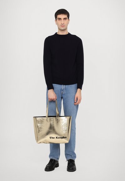 Zlatna metalik torba s crnim unutrašnjim djelom i logom "The Kooples", koju drži osoba u tamnoplavoj majici i plavim trapericama.