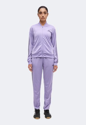 Frau steht mit Blick nach vorne, trägt einen passenden hellvioletten Adidas-Trainingsanzug mit schwarzen Sneakers vor einem schlichten Hintergrund.
