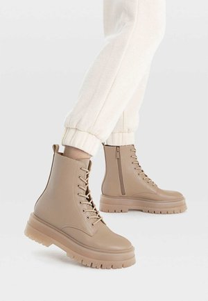 Beige chunky snørestøvler til anklen med sidelås, båret med hvide joggerbukser, vist fra siden på en hvid baggrund.