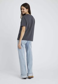 T-shirt en coton gris à manches courtes, coupe décontractée. Associé à un jean bleu clair à jambe large. Le mannequin est légèrement tourné, montrant le dos.