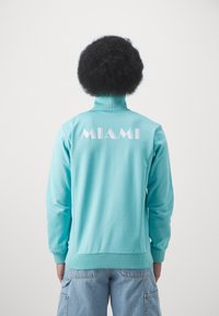 Lys blå zip-up jakke med høj krave, der har ordet "MIAMI" i hvide geometriske bogstaver på ryggen. Glat tekstur, ribbede manchetter.