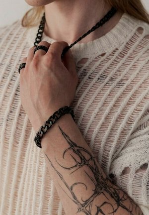 Person trägt einen weißen offen gestrickten Pullover, hält eine schwarze Kettenkette, mit schwarzen Ringen, Armband und einem detaillierten schwarzen Tattoo auf dem Unterarm.