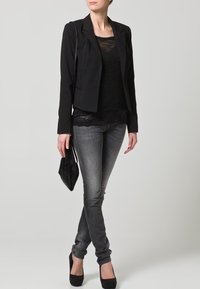 Schwarzer Blazer über einem schwarzen Oberteil mit Spitzenbesatz, kombiniert mit grauen Skinny-Jeans und schwarzen Plateau-Heels. Hält eine kleine schwarze Clutch.