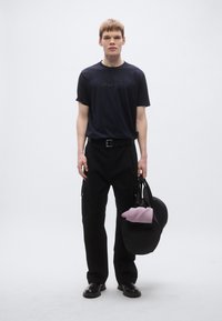 Fekete rövid ujjú póló "BOSS" logóval, fekete nadrággal oldalzsebekkel, és fekete cipővel. Egy fekete táskát tart lila részlettel.