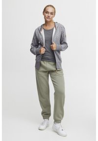 Grå zip-up hoodie med spräcklig mönster, vit foder och sidofickor, kombinerad med ljusgröna träningsoverallsbyxor och vita sneakers.