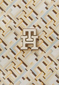 Hvid og beige mønstret stof med geometrisk design, der har et fremtrædende logo-emblem i guld og creme i midten.