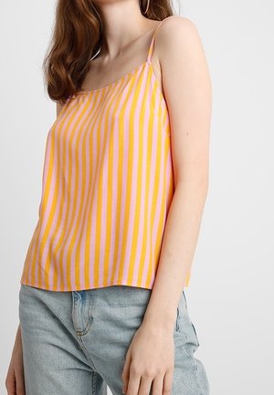 Frau in einem ärmellosen, pink- und gelbgestreiften Oberteil, kombiniert mit hellblauen Jeans, steht vor einem schlichten Hintergrund.