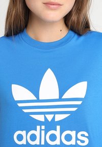 Modré bavlněné tričko s velkým bílým logem Adidas, které obsahuje tři listovité tvary a dvě vodorovné pruhy pod textem.