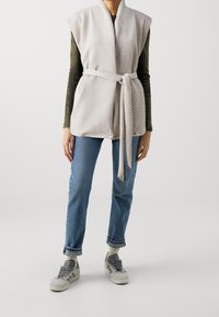 Světlá šedá vesta s diagonálním žebrovaným vzorem, páskem v oblasti pasu, otevřeným předním dílem a krátkými rukávy, nošená přes tmavě zelený top s dlouhým rukávem a modré džíny.