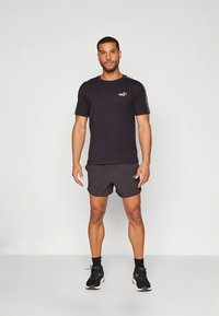 Črna kratka športna majica s kratkimi rokavi z logotipom, v kombinaciji z sivimi športnimi kratkimi hlačami. Model nosi črne in bež superge. Enostavna, športna zasnova.