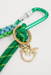 Grünes und blaues geflochtenes Band mit einem grünen Karabiner, einem goldenen Ring und zwei Vogelanhängern. Verfügt über eine kunstvolle Textur und kontrastierende Farben.