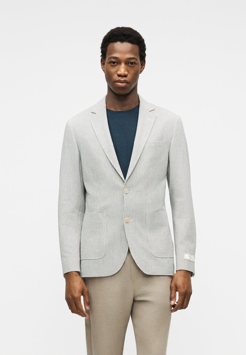 Mand iført en lysegrå tekstureret blazer over en mørkeblå skjorte og beige bukser, stående mod en ensartet lys baggrund.