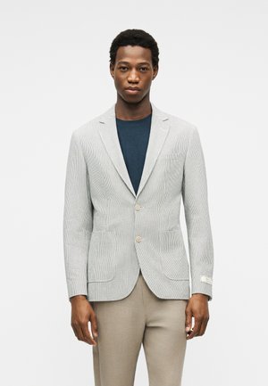Mand iført en lysegrå tekstureret blazer over en mørkeblå skjorte og beige bukser, stående mod en ensartet lys baggrund.
