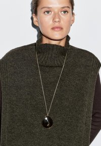 Dark green knitted turtleneck sweater with textured ribbing and short sleeves, paired with a long gold chain necklace featuring a circular pendant.