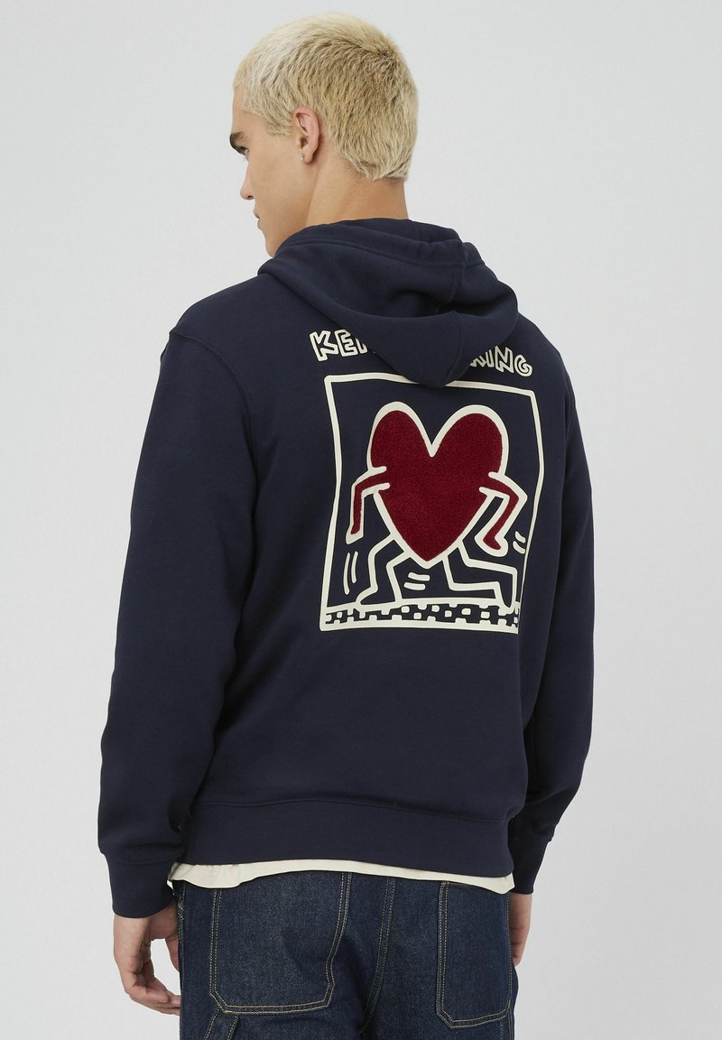 Sweat à capuche bleu marine arborant un grand motif de cœur rouge texturé au dos, entouré de crème. Il a une coupe décontractée et une poche kangourou.