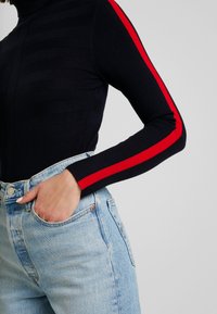Zwarte geribbelde coltrui met rode strepen langs de mouwen, gecombineerd met lichtblauwe high waist jeans. Soepele stof en aangesloten ontwerp.