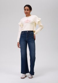 Felpa cream a maniche a campana con volant, decorata con una grafica della Torre Eiffel rosa, abbinata a jeans svasati blu scuro e sneakers neutre.