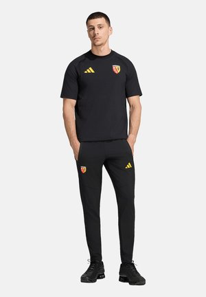 Mężczyzna stoi z rękami w kieszeniach, ubrany w czarą odzież sportową z żółtymi logo Adidas i czerwono-żółtym herbem RCL na koszulce i spodniach.