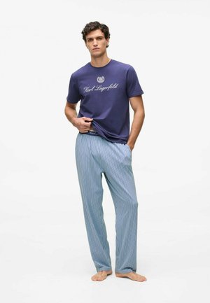 Jeune homme portant un t-shirt bleu Karl Lagerfeld et un pantalon décontracté bleu clair à rayures verticales, debout pieds nus sur un fond blanc uni.