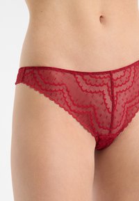 Braguitas hipster de encaje rojo con bordes ondulados, patrones intrincados y una textura ligera y translúcida.