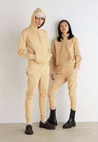 Sudaderas con capucha beige y pantalones a juego, de mezcla de algodón, con logotipo bordado. Un modelo lleva una gorra y el otro, botas negras.