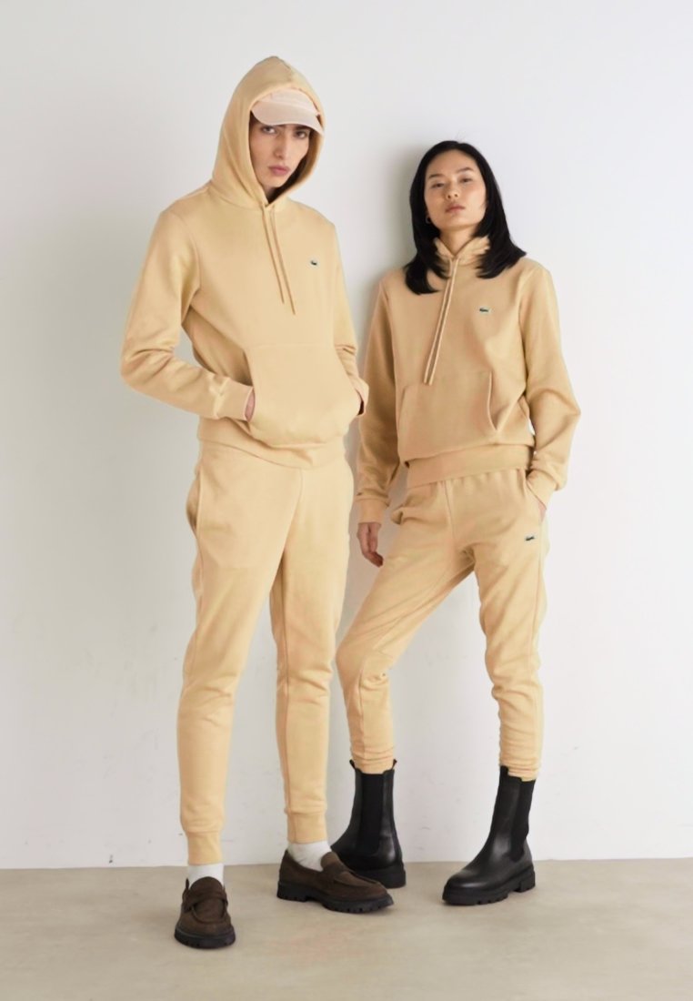 Sweatshirts à capuche beige et pantalons de jogging assortis, en mélange de coton, avec logo brodé. Un modèle porte une casquette et l'autre, des bottes noires.