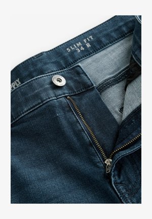Jeans de denim ajustados en azul oscuro; cuentan con un cierre de metal, botonera y tela interior en contraste. La etiqueta muestra la talla 34 R.