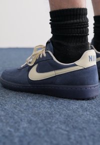 Baskets en suède bleu marine avec un swoosh et un logo de couleur crème, dotées de lacets crème et d'une semelle en caoutchouc texturée.