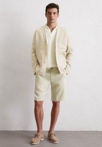 Beige Leinenblazer, hellgrüne Shorts, strukturiertes helles Polo-Shirt, braune Schuhe. Einfaches Design, lässige Passform, keine auffälligen Muster, neutrale Farben.