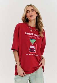 Czerwona, oversize'owa lekka koszulka z miękkiego materiału, z grafiką dzbanka do kawy i napisem w białym i różowym kolorze. Krótkie rękawy i luźny krój.
