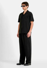 Chemise noire à manches courtes avec un bouton, portée avec un pantalon noir ample et des chaussures noires épaisses, créant un ensemble monochrome.