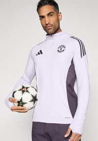 Λευκό αθλητικό πουκάμισο μακρυμάνικο με γιακά μισού φερμουάρ, σκουρόχρωμες γκρι πλευρικές πινακίδες και λογότυπο της Μάντσεστερ Γιουνάιτεντ. Κρατάει μια μπάλα ποδοσφαίρου.