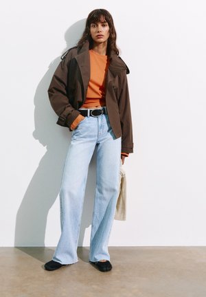 Femme debout contre un mur blanc, portant une veste marron, un haut orange, un jean bleu clair, des chaussures noires, et tenant un sac beige.